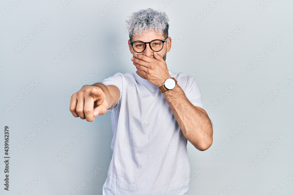 Young hispanic man with modern dyed hair wearing white t shirt and glasses laughing at you, pointing finger to the camera with hand over mouth, shame expression Young hispanic man with modern dyed hair wearing white t shirt and glasses laughing at you, pointing finger to the camera with hand over mouth, shame expression