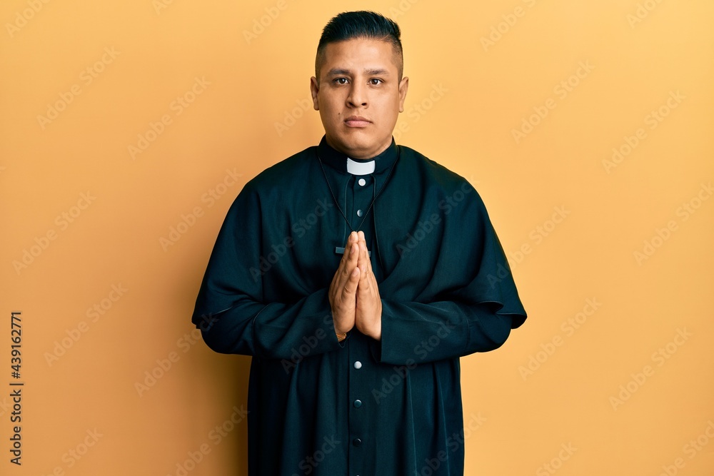 Young latin priest man praying with hands together relaxed with serious expression on face. simple and natural looking at the camera. Young latin priest man praying with hands together relaxed with serious expression on face. simple and natural looking at the camera.