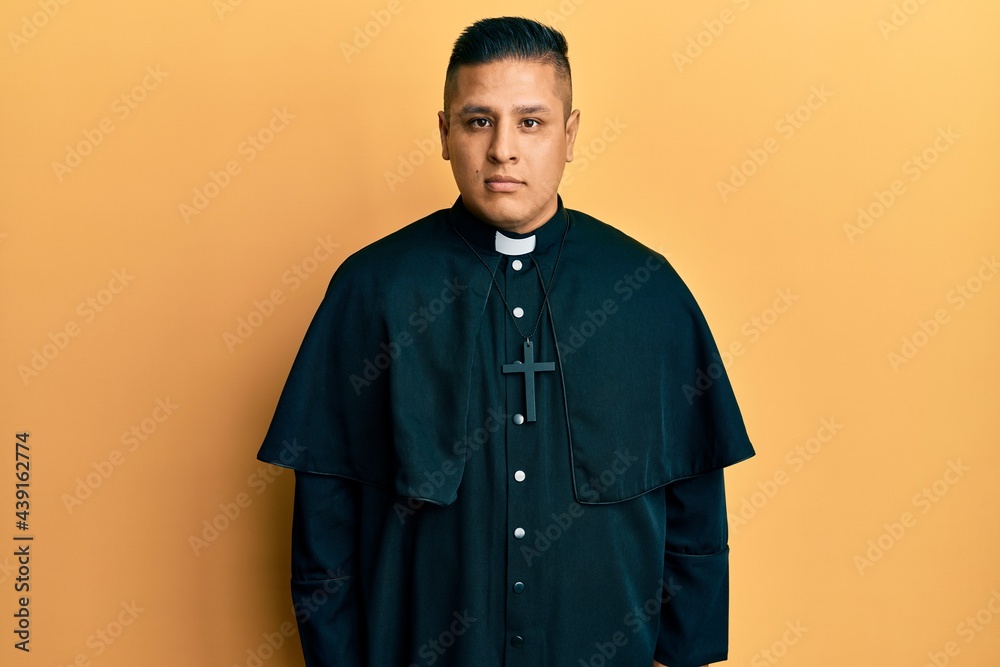 Obraz premium Young latin priest man standing over yellow background relaxed with serious expression on face. simple and natural looking at the camera.
