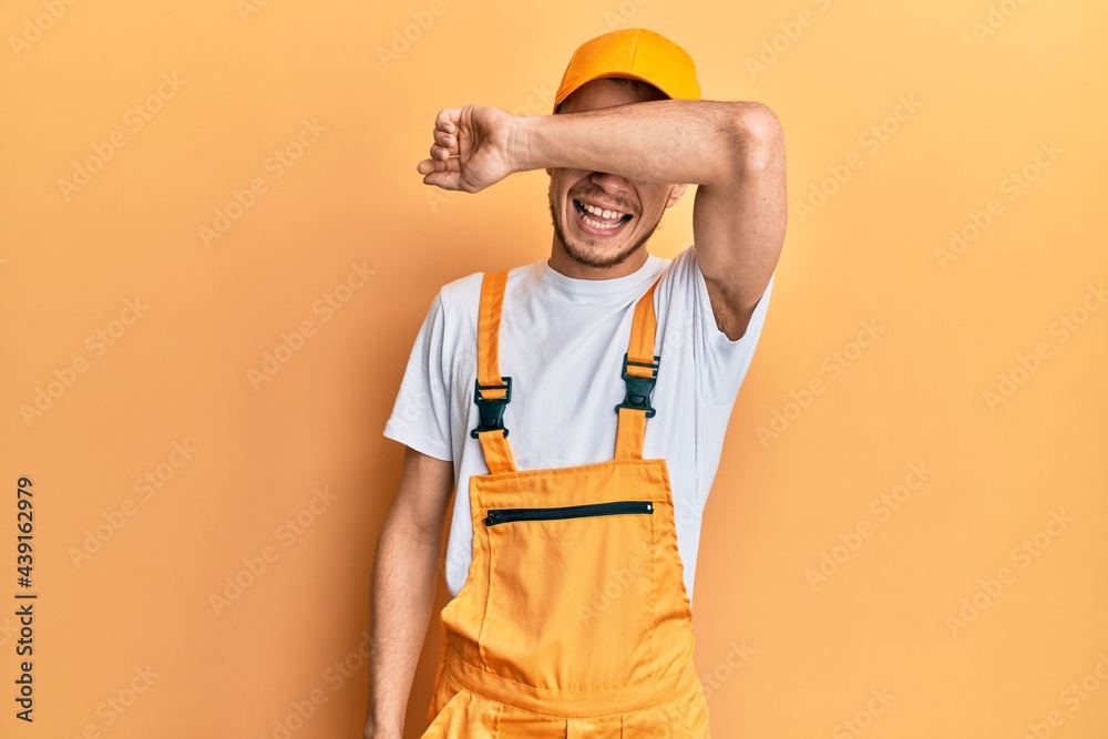 Hispanic young man wearing handyman uniform covering eyes with arm smiling cheerful and funny. blind concept. Hispanic young man wearing handyman uniform covering eyes with arm smiling cheerful and funny. blind concept.