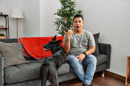 Young latin man and dog sitting on the sofa at home surprised pointing with hand finger to the side, open mouth amazed expression. © Krakenimages.com