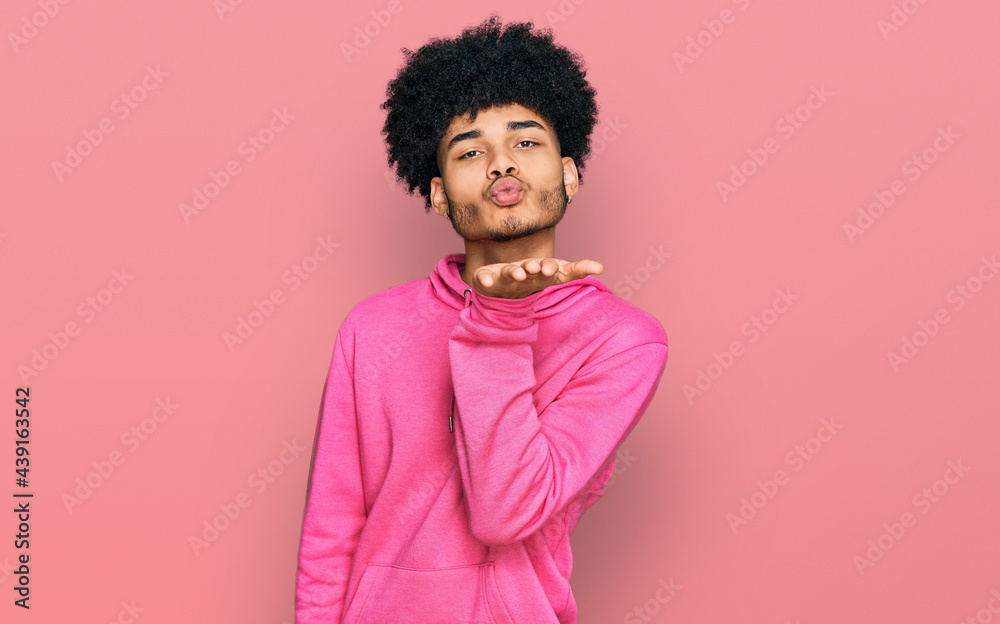 Young african american man with afro hair wearing casual pink sweatshirt looking at the camera blowing a kiss with hand on air being lovely and sexy. love expression. Young african american man with afro hair wearing casual pink sweatshirt looking at the camera blowing a kiss with hand on air being lovely and sexy. love expression.