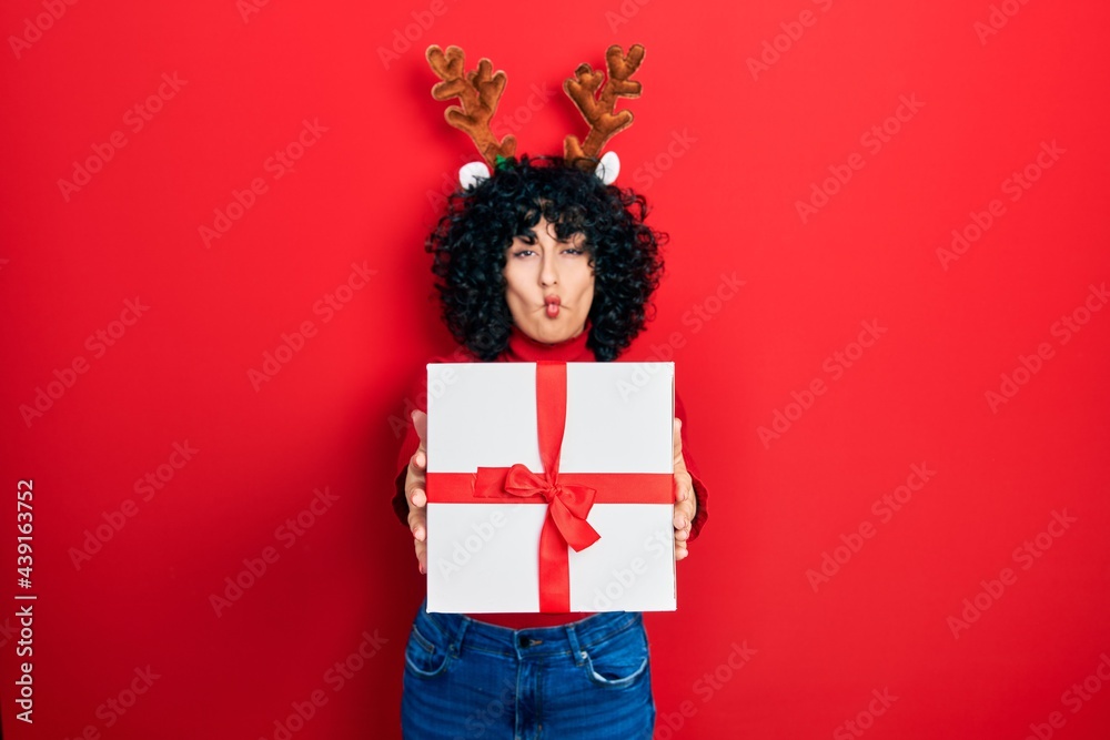 Fototapeta premium Young middle east woman wearing deer christmas hat holding gift making fish face with mouth and squinting eyes, crazy and comical.