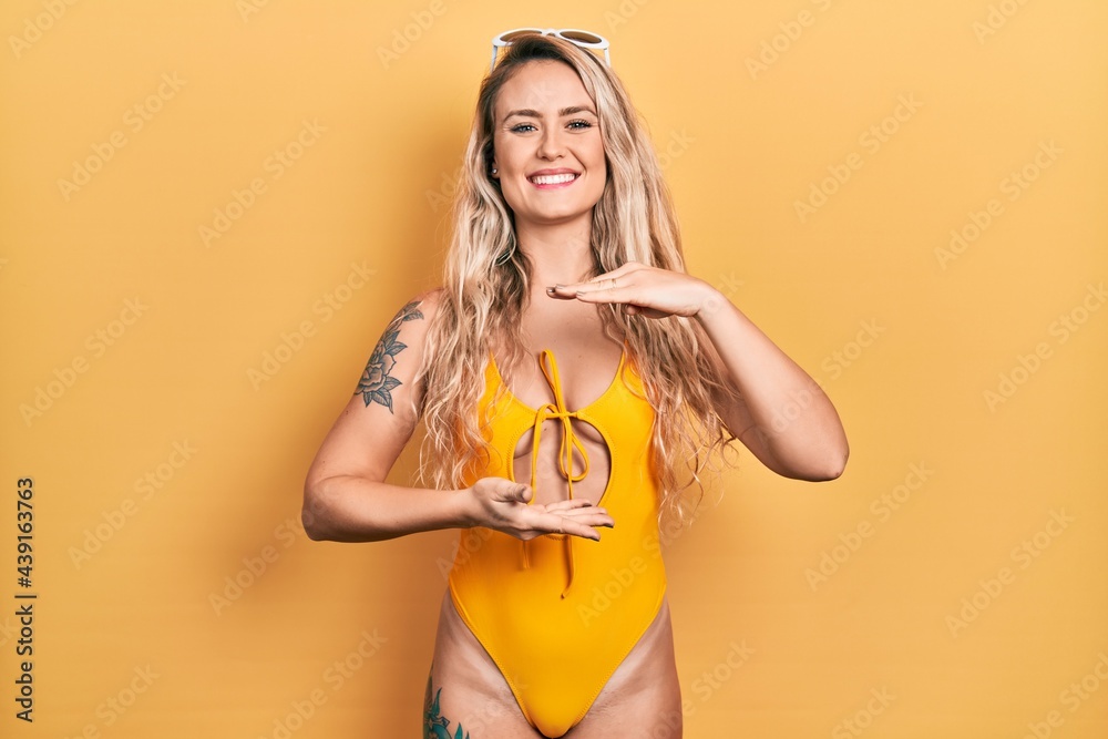 Beautiful young blonde woman wearing swimsuit and sunglasses gesturing with hands showing big and large size sign, measure symbol. smiling looking at the camera. measuring concept. Beautiful young blonde woman wearing swimsuit and sunglasses gesturing with hands showing big and large size sign, measure symbol. smiling looking at the camera. measuring concept.