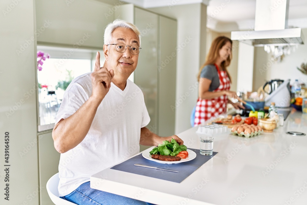 Middle age hispanic couple eating healthy meal at home surprised with an idea or question pointing finger with happy face, number one Middle age hispanic couple eating healthy meal at home surprised with an idea or question pointing finger with happy face, number one