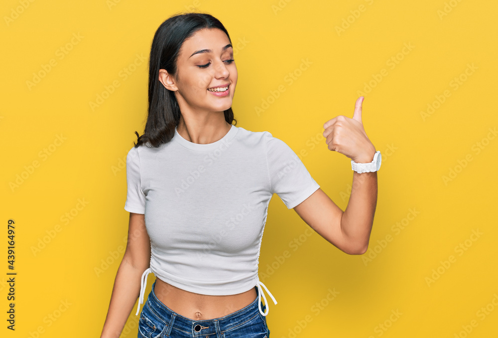 Young hispanic girl wearing casual white t shirt looking proud, smiling doing thumbs up gesture to the side Young hispanic girl wearing casual white t shirt looking proud, smiling doing thumbs up gesture to the side