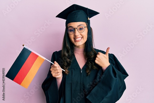 Young hispanic woman wearing graduation uniform holding germany flag smiling happy and positive, thumb up doing excellent and approval sign