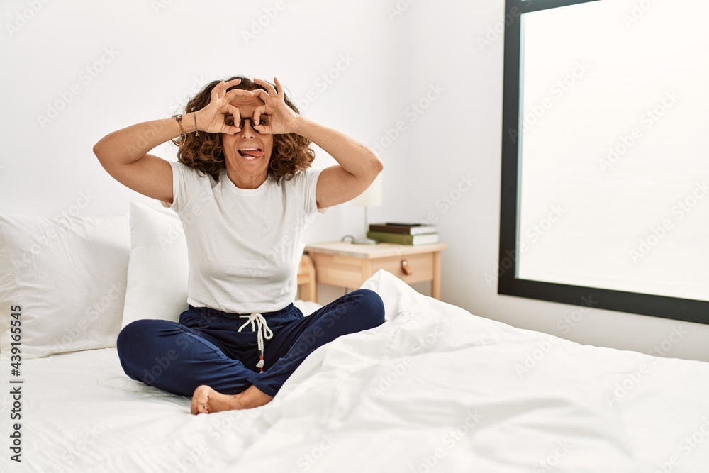 Obraz premium Middle age hispanic woman sitting on the bed at home doing ok gesture like binoculars sticking tongue out, eyes looking through fingers. crazy expression.