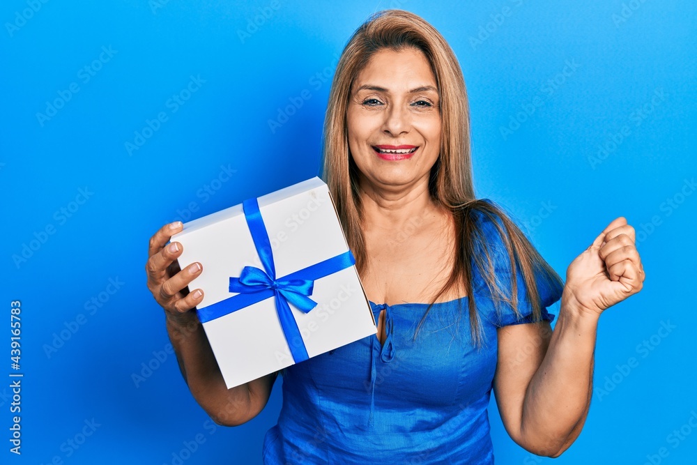Middle age hispanic woman holding gift screaming proud, celebrating victory and success very excited with raised arm Middle age hispanic woman holding gift screaming proud, celebrating victory and success very excited with raised arm