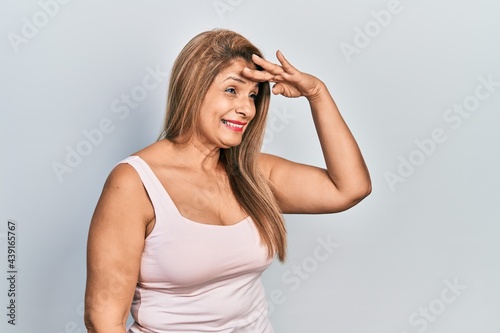 Middle age hispanic woman wearing casual style with sleeveless shirt very happy and smiling looking far away with hand over head. searching concept.