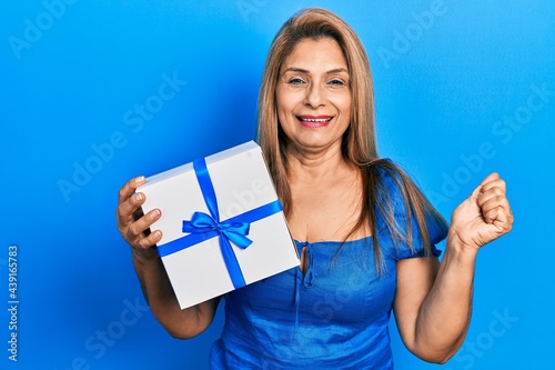 Middle age hispanic woman holding gift screaming proud, celebrating victory and success very excited with raised arm