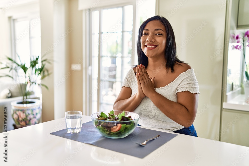 Young hispanic woman eating healthy salad at home praying with hands together asking for forgiveness smiling confident.