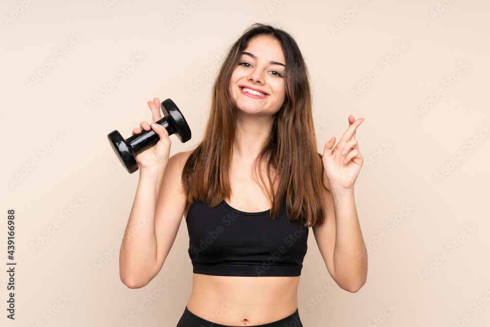 Young sport woman making weightlifting isolated on beige background with fingers crossing Young sport woman making weightlifting isolated on beige background with fingers crossing