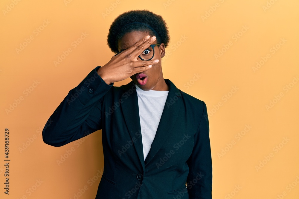 Young african american girl wearing business jacket and glasses peeking in shock covering face and eyes with hand, looking through fingers afraid