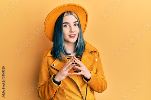 Young modern girl wearing yellow hat and leather jacket hands together and fingers crossed smiling relaxed and cheerful. success and optimistic
