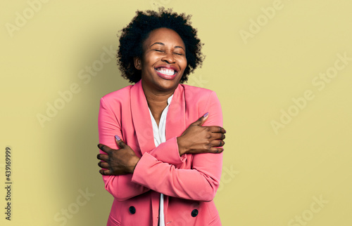 African american woman with afro hair wearing business jacket hugging oneself happy and positive, smiling confident. self love and self care
