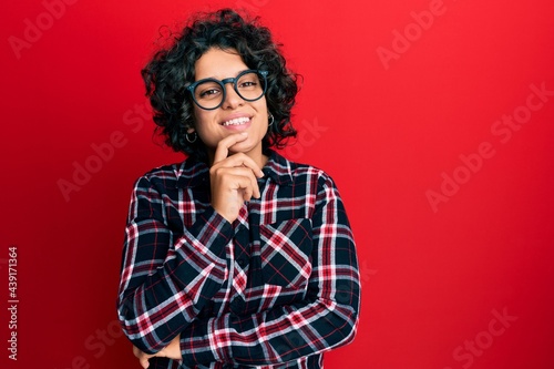 Young hispanic woman with curly hair wearing casual clothes and glasses looking confident at the camera with smile with crossed arms and hand raised on chin. thinking positive.