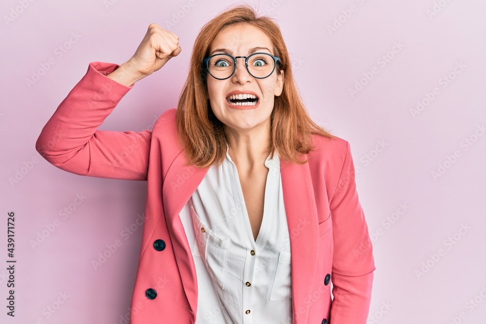 Young caucasian woman wearing business style and glasses angry and mad raising fist frustrated and furious while shouting with anger. rage and aggressive concept.