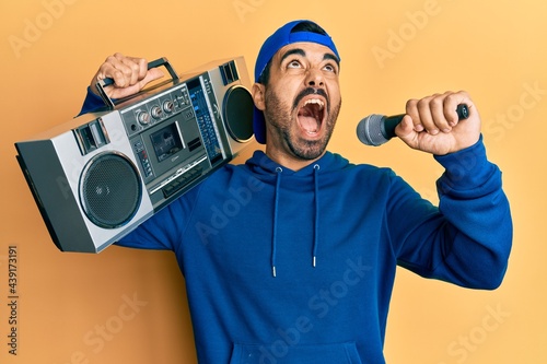 Young hispanic man holding boombox, listening to music singing with microphone angry and mad screaming frustrated and furious, shouting with anger looking up.