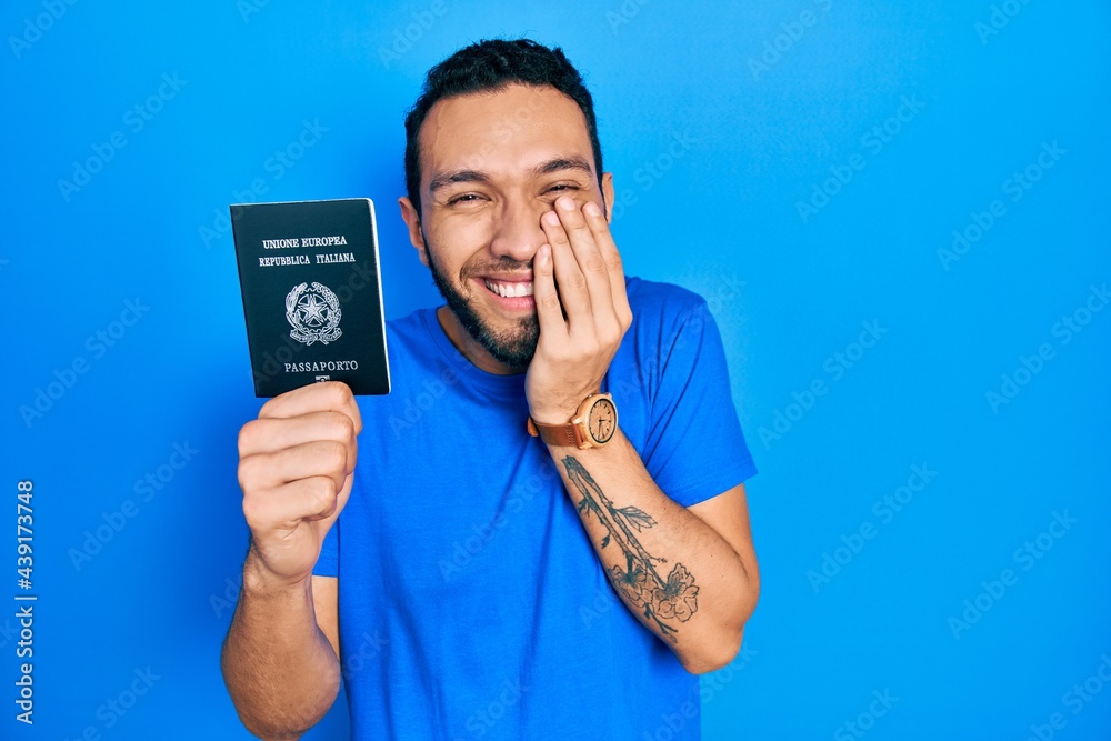 Hispanic man with beard holding italy passport laughing and embarrassed giggle covering mouth with hands, gossip and scandal concept Hispanic man with beard holding italy passport laughing and embarrassed giggle covering mouth with hands, gossip and scandal concept