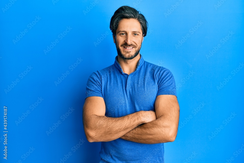 Young hispanic man wearing casual blue t shirt happy face smiling with crossed arms looking at the camera. positive person. Young hispanic man wearing casual blue t shirt happy face smiling with crossed arms looking at the camera. positive person.