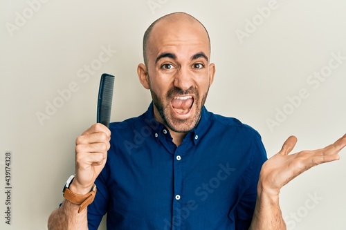 Young hispanic man holding comb loosing hair celebrating victory with happy smile and winner expression with raised hands