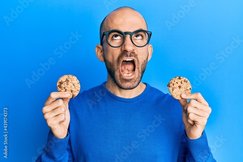 Young hispanic man holding chocolate chips cookies angry and mad screaming frustrated and furious, shouting with anger looking up Fotobehang