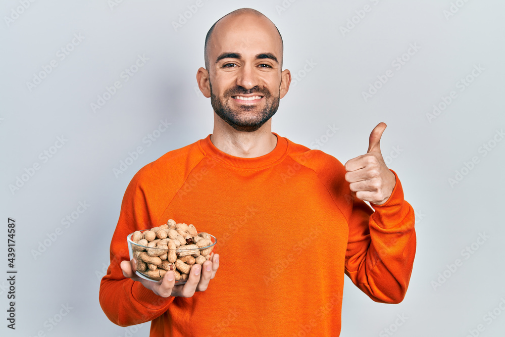 Young bald man holding peanuts smiling happy and positive, thumb up doing excellent and approval sign Young bald man holding peanuts smiling happy and positive, thumb up doing excellent and approval sign