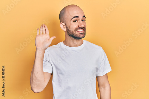 Young bald man wearing casual white t shirt waiving saying hello happy and smiling, friendly welcome gesture