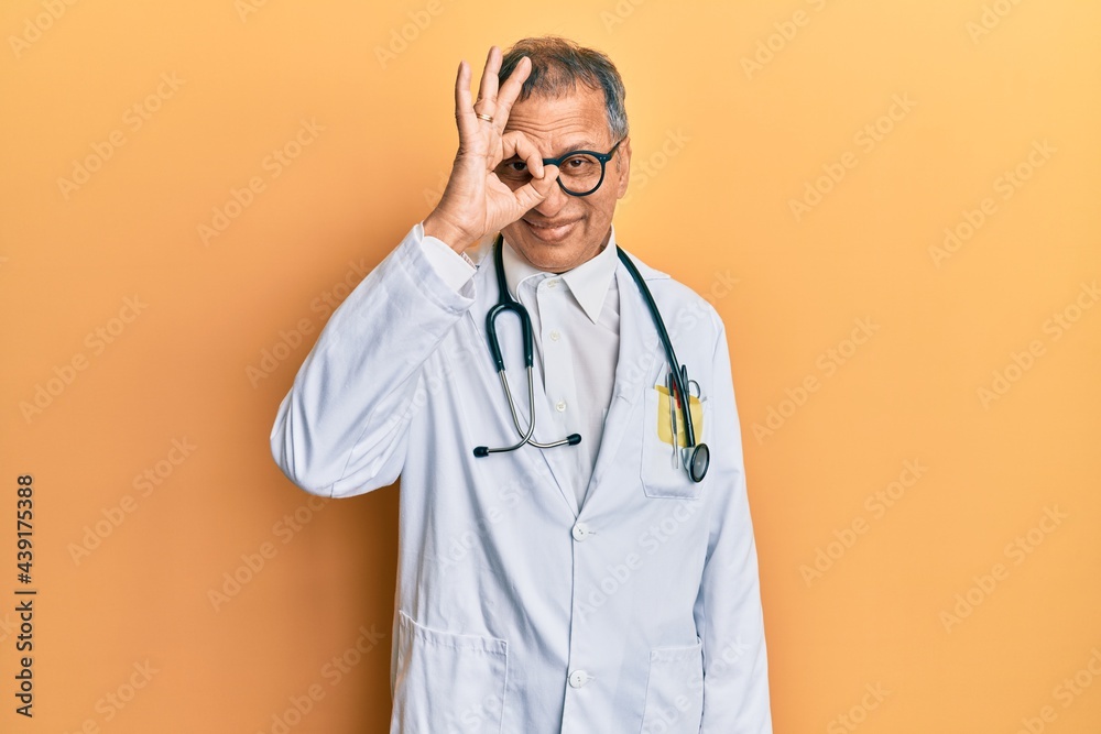 Middle age indian man wearing doctor coat and stethoscope doing ok gesture with hand smiling, eye looking through fingers with happy face. Middle age indian man wearing doctor coat and stethoscope doing ok gesture with hand smiling, eye looking through fingers with happy face.