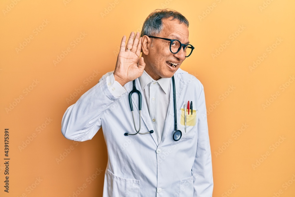 Middle age indian man wearing doctor coat and stethoscope smiling with hand over ear listening an hearing to rumor or gossip. deafness concept.