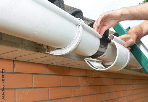 Roof gutter installation and repair. A man on a ladder is replacing a plastic rain gutter joint, bracket, connecting the gutters together. photo