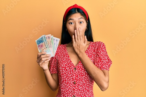 Photography Young chinese woman holding singapore dollars banknotes covering mouth with hand, shocked and afraid for mistake