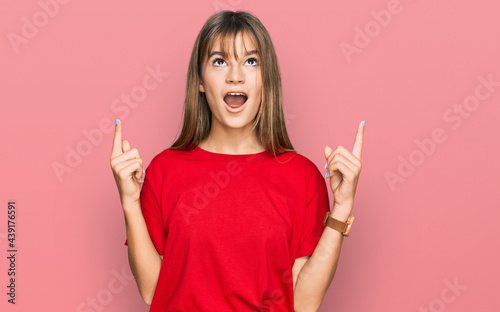 Teenager caucasian girl wearing casual red t shirt amazed and surprised looking up and pointing with fingers and raised arms.