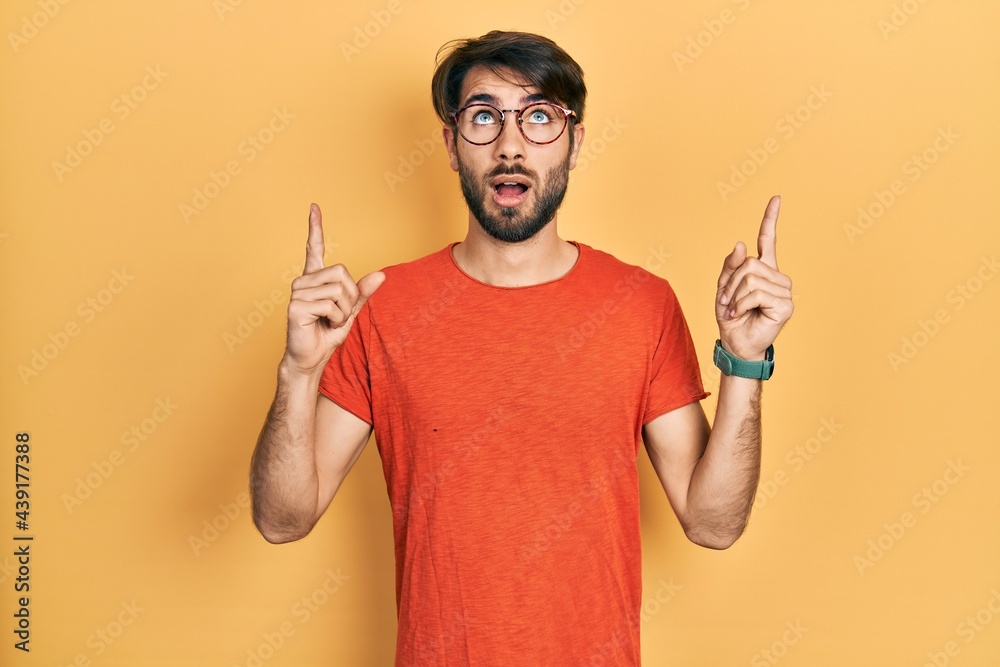 Young hispanic man wearing casual clothes and glasses amazed and surprised looking up and pointing with fingers and raised arms.