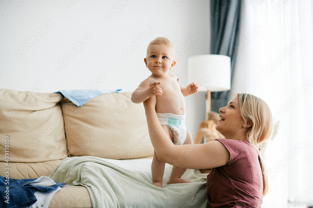 Cute baby boy in diapers standing on the sofa while mother is holding him.