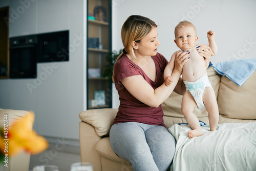 Baby boy standing on the sofa with help of his mother at home.