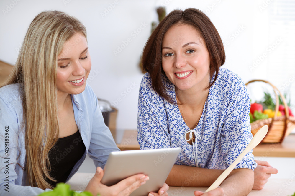 Two women friends choosing the recipe for a delicious meal while sitting at the table in the kitchen. Tablet pc is the best cookbook Two women friends choosing the recipe for a delicious meal while sitting at the table in the kitchen. Tablet pc is the best cookbook