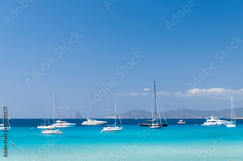 Various sailboats moored in the turquoise sea near a beach on the island of Formentera. In the background the blue sky and the coast of Ibiza
