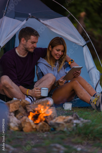 Young couple drinking coffee, enjoying the mountain view and checking the tablet with bonfire in the sunset