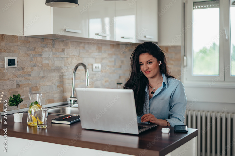 Woman using a laptop and airpods while sitting in the kitchen Woman using a laptop and airpods while sitting in the kitchen