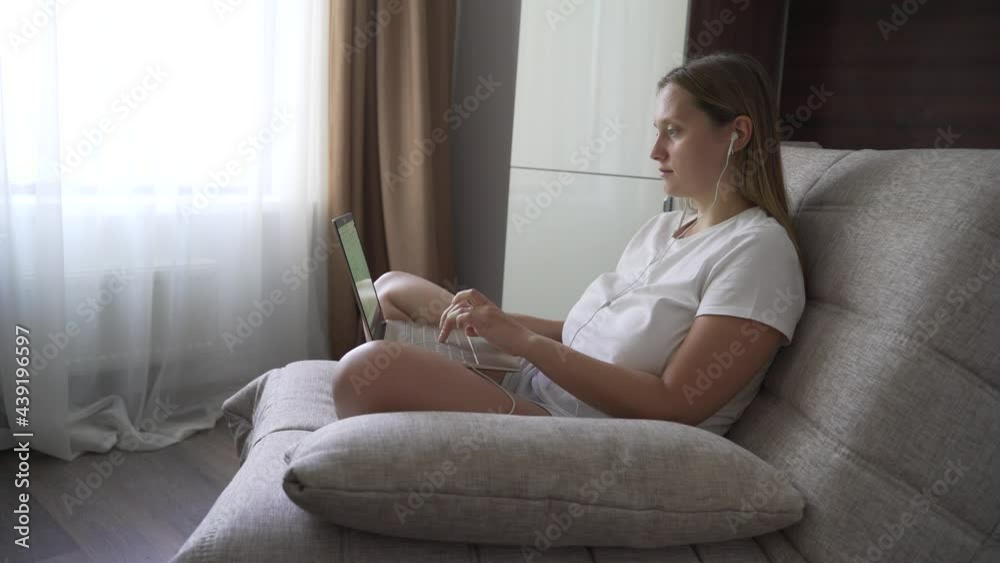 Woman in white T-shirt sits on armchair near the window in headphones and works on notebook in the early morning alone at home in quarantine. Typing text on the keyboard on the computer Woman in white T-shirt sits on armchair near the window in headphones and works on notebook in the early morning alone at home in quarantine. Typing text on the keyboard on the computer