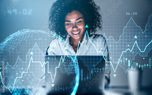 Smiling African-American businesswoman working on laptop researching financial news. Stock market changes candlesticks, data information icons. Globe projection. Concept of finance and technology