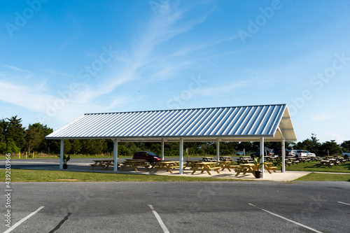 Wooden crab boil picnic feast tables gathered underneath a newly built smart shelter pavilion with solar panel collector roof from composite materials seen from the parking lot.