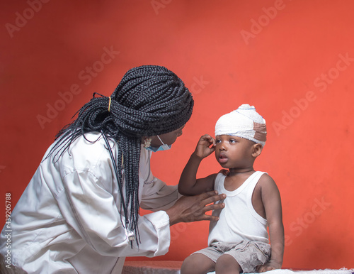 African female nurse, doctor, or medical specialist with nose mask and stethoscope doing health checks and treatment on a little boy child that has wound bandage wrapped around his head