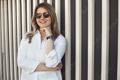 Lifestyle portrait of a smiling young confident woman 30-40 years old in a white shirt, wears sunglasses and a wrist watch