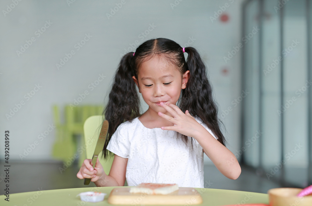 Portrait happy little child girl cooking at class room. Kid taste strawberry jam while cooking. Children are using knife slicing and apply jam on bread on chopping wood board. Portrait happy little child girl cooking at class room. Kid taste strawberry jam while cooking. Children are using knife slicing and apply jam on bread on chopping wood board.