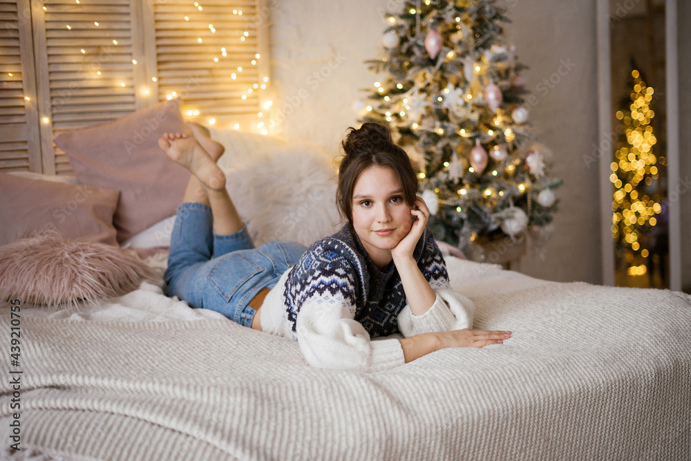 A young woman lies on a bed in a bright bedroom. Christmas cozy interior with a Christmas tree and garlands in the background. A young woman lies on a bed in a bright bedroom. Christmas cozy interior with a Christmas tree and garlands in the background.