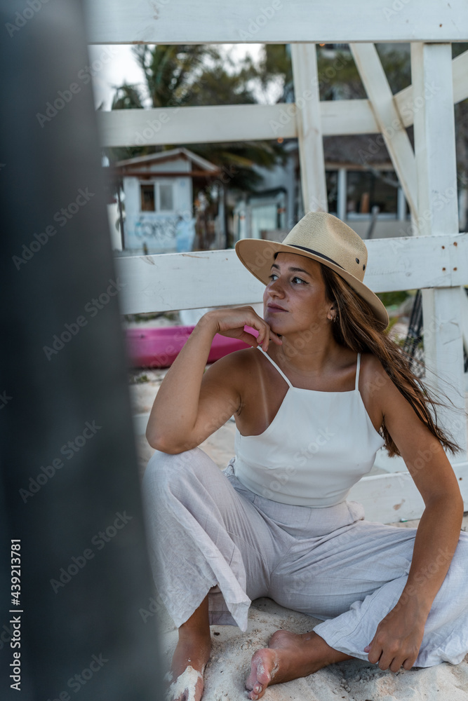 young hispanic woman with hat posing sitting on the sand looking at the sea - latinx