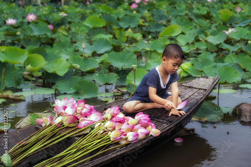 Obraz na płótnie Vietnamese boy playing with pink lotus leaf when his mom boating the traditional wooden boat in the big lake at thap muoi, dong thap province, vietnam, culture and life concept w salonie